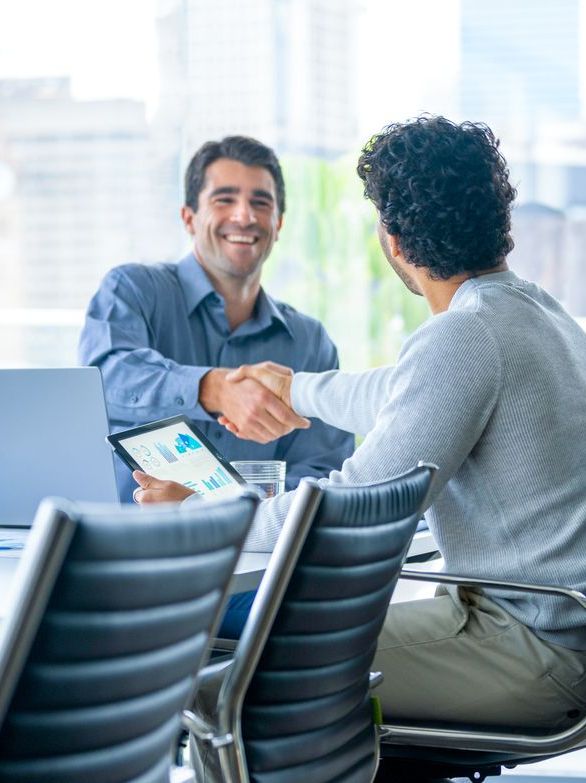 Two businessmen working on a digital tablet and laptop computer in a boardroom or meeting room.